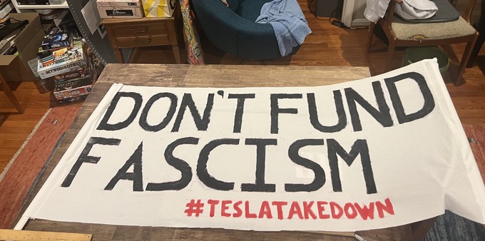 Photo of a banner laying across a table. It’s about four feet long and 2 feet tall. It says in big black letters “don’t fund fascism” and the hashtag tesla take down in red