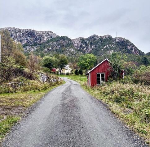 This image features a rural landscape dominated by lush greenery and a rugged mountain background. A gravel road curves through the scene, leading past a small, traditional red wooden shed on the left and onward to a cluster of houses. There's a two-story yellow house visible in the distance that adds a quaint, residential feel to the otherwise natural setting. The overall vibe is serene and picturesque, typical of Scandinavian countryside environments.