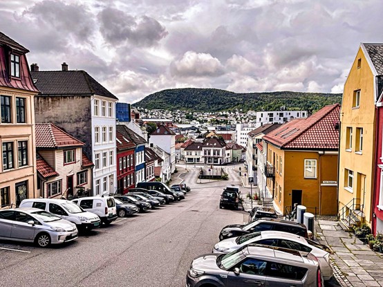 The image depicts a picturesque urban scene, possibly in a European town, characterized by quaint, narrow streets flanked by traditional houses with distinctively colorful facades. The scene includes parked cars lining the street, suggesting a residential area. In the background, there are more modern buildings and a hilly landscape, indicating that the town is located in a valley or near hills. The overall cloudy sky contributes to a serene atmosphere typical of a temperate climate.