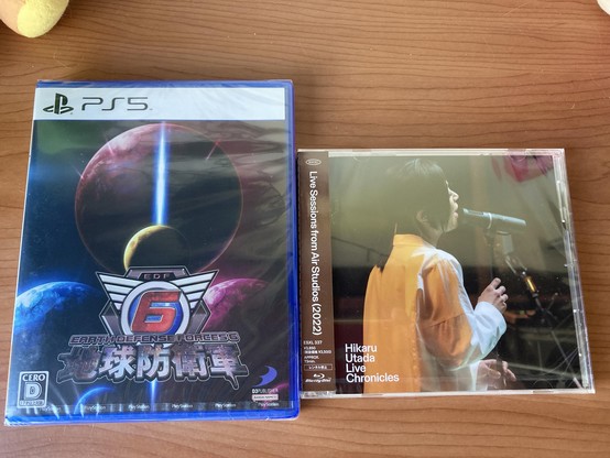 The image features two entertainment items: a PlayStation 5 game titled "Earth Defense Forces 6" with vibrant planetary graphics, and a CD titled "Hikaru Utada Live Chronicles," showcasing a performer singing into a microphone.