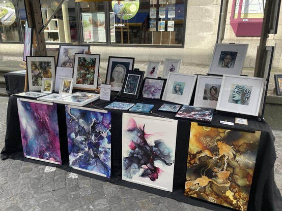 Eine Ausstellung mit verschiedenen Kunstwerken, darunter farbenfrohe abstrakte Gemälde und gerahmte Porträts. Das Kunstwerk zeigt eine Reihe von Stilen und leuchtenden Farben, die auf einem mit einem schwarzen Tuch bedeckten Tisch in einer Marktumgebung im Freien angeordnet sind.