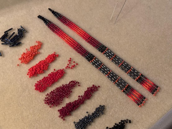 A pair of long beaded dangles in shades of red, grey, and black, on a bead mat next to piles of seed beads in the same colors. A few needles stick out of the bead mat.