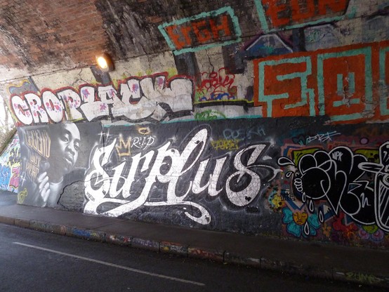 In a tunnel, light coming from the entrance to left is reflected in the silver letters Sirplus, and a portrait of a Black Bristolian rapper