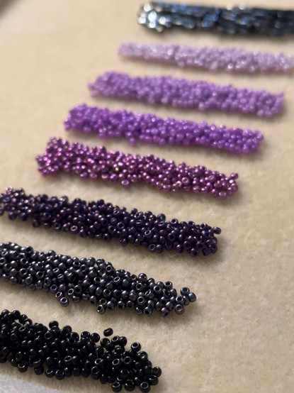 A line of seed bead piles in shades of purple, grey, and black, on a cream colored bead mat