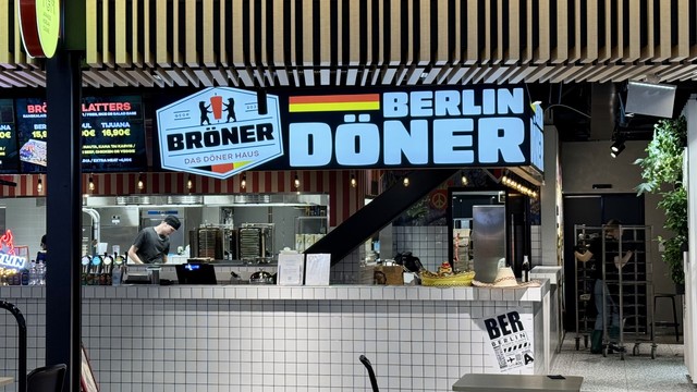 Image of a food stall named "Brönner - Das Döner Haus," featuring bright signage that includes "Berlin Döner." A staff member in a black shirt and hat is seen working behind the counter. The menu displays various food options and prices.