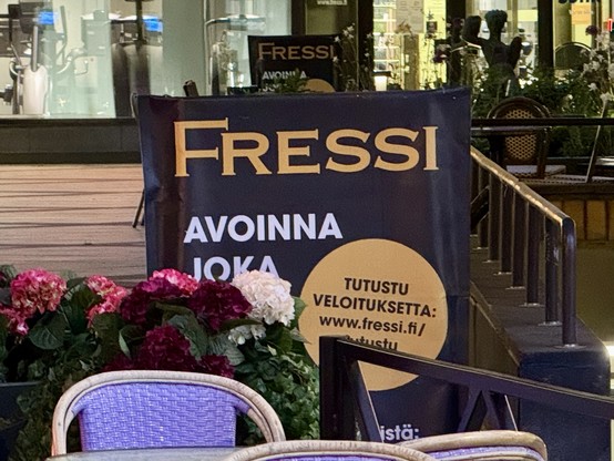 A promotional banner for "Fressi" with text indicating it's open every day. There are decorative flowers in front and some outdoor furniture visible. The background shows a well-lit indoor setting.