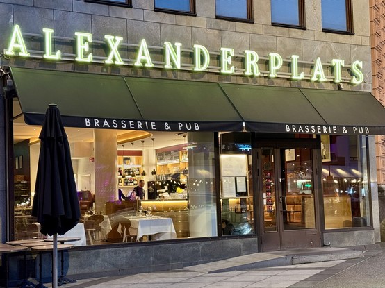 A restaurant and pub named "Alexanderplatz" featuring a stylish exterior with illuminated signage. The entrance is adorned with black awnings and a table with an umbrella, while the interior is visible through large windows, showcasing a bar area and seating.