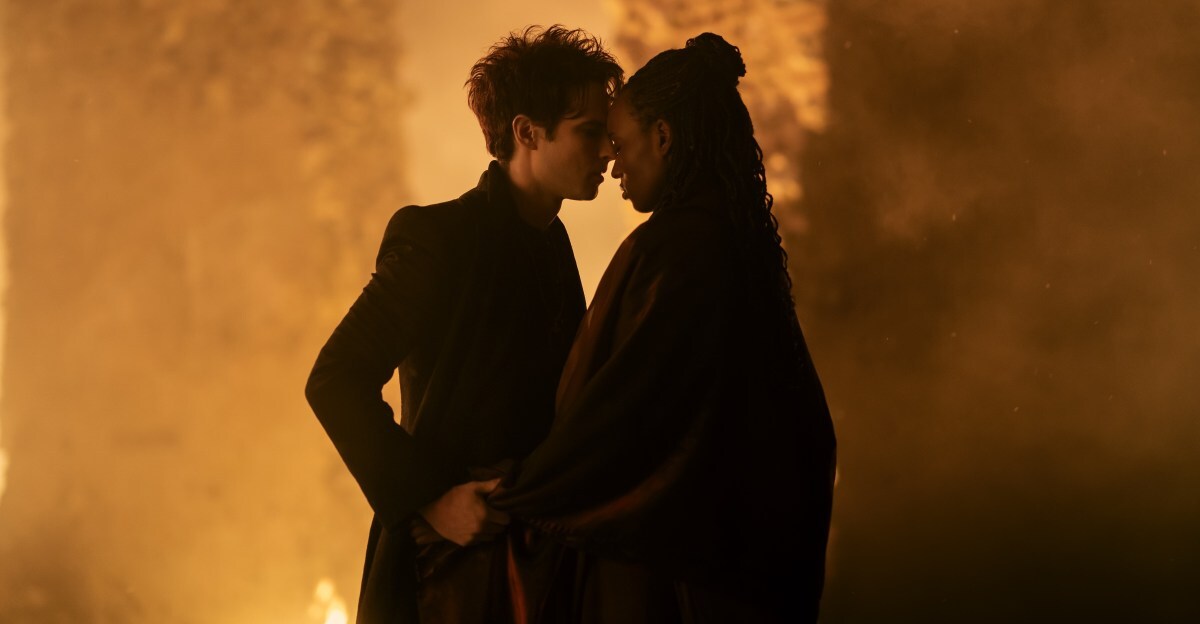 A man in a black suit embracing a woman in a black dress and cape. The couple are backlit by an orange, glowing light.