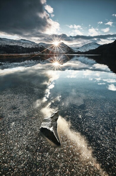 The sun sparkles as it sets behind the hills around Llyn Dinas in Eryri. It is winter, snow lies on the mountains. The water is calm and the landscape is reflected in it. 