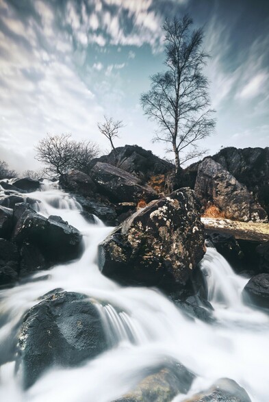 The Idwal River in Eryri flows fast, filled with melting winter snow. 
A long exposure smooths out the water around the rocks. The tree above is bare.