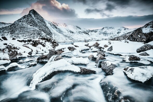 Looking the valley of Dyffryn Ogwen at sunset towards Tryfan and the mountains beyond. It is winter and the landscape is covered with snow and ice. The stream in the foreground is starting to freeze over.