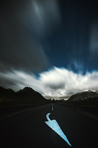 A long exposure in the middle of a road at night in Eryri. The clouds move quickly above and the stars are revealed. Mountains to the left and right. 

A road sign points to the bottom of the frame. 
