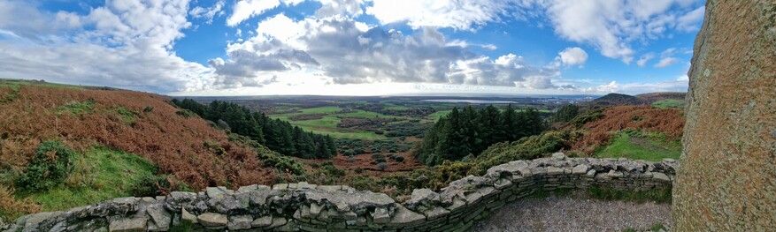 Panoramic view , The edge of a large standing stone can be seen to the right of the picture. A low  stone wall in the foreground, looking down over green hills covered in bushes and some trees , green farmland in the distance and the sea in the far distance. Sky is patches of blue but mostly light white or grey clouds