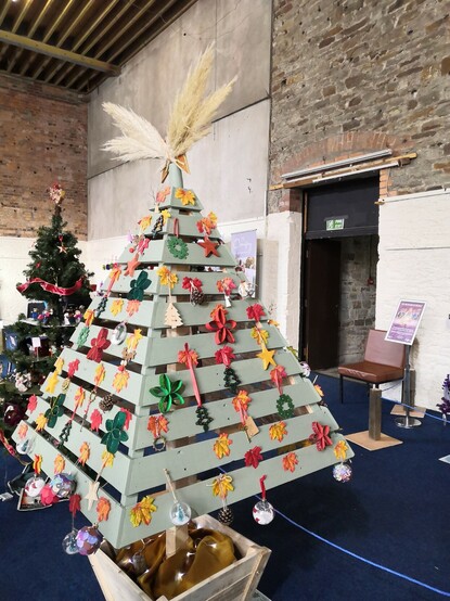 Four-sided pyramid tree, made from strips of recycled wood, with decorations pinned to the strips in rows.