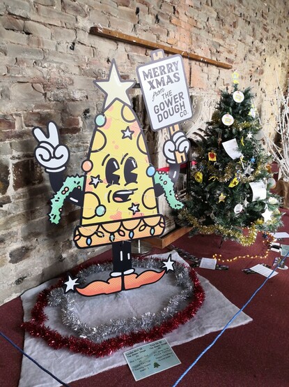 Pizza Christmas tree from Gower Dough Co., a cartoon figure of a smiling pizza holding up a sign and making a 'peace' hand gesture.