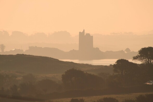 a view over fields and a loch to a old but renovated tower/castle built on a small island. The Castle is shrouded in an atmospheric early morning mist giving the picture a warm orange tint