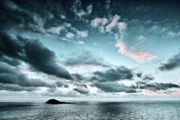 Looking out across the Irish Sea towards Ynys Enlli. It is sunset. The sky is filled with dramatic clouds as the last of the warm sunset colours begin to fade. 

The island sits isolated on a silvery sea.