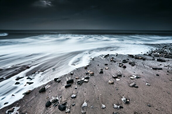 A smooth, velvet-like wave washes onto the pebbly beach at Dinas Dinlle. The sky beyond is dark and brooding, bad, winter weather is on the way.