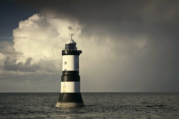 The lighthouse at Trwyn Du, Penmon, Ynys Môn. 

The lighthouse sits alone on a calm sea. Sunlight comes into frame from the left. Behind the lighthouse the weather looks dark and full of drama.
