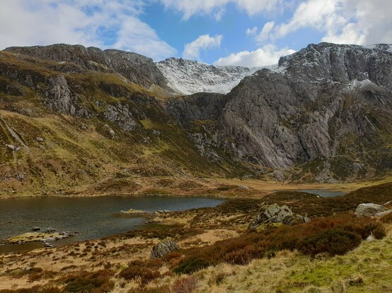 A still lake in the middle of green and brown heathland with dramatic mountains rising up behind.The highest slopes of the mountains have a light dusting of snow. The sky is blue with some white whispy clouds 
