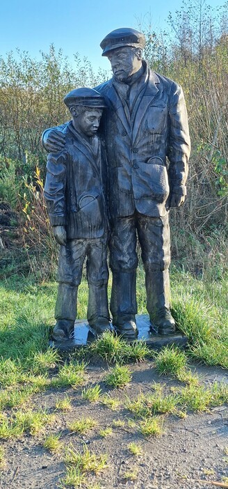 An almost life size wooden carving depicting two figures probably on their way to work in a coal mine. A man with his arm around the shoulders of a boy, both wearing loose jackets and trousers, working boots and flat caps