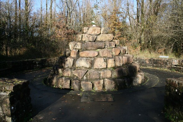 A monument in the shape of a circular pyramid built of large rough hewn stones . A low wall surrounds the monument with an opening for people to approach and walk around, A plaque is set in the floor, but the writing cannot be seen in the picture.  Cut flowers have been placed on the top of the pyramid