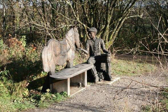 An almost life size wooden carving of a young man, dressed in work clothes , sitting and feeding a pit pony by hand from a bag on his knee. A wooden bench extends from the pony forming part of the carving