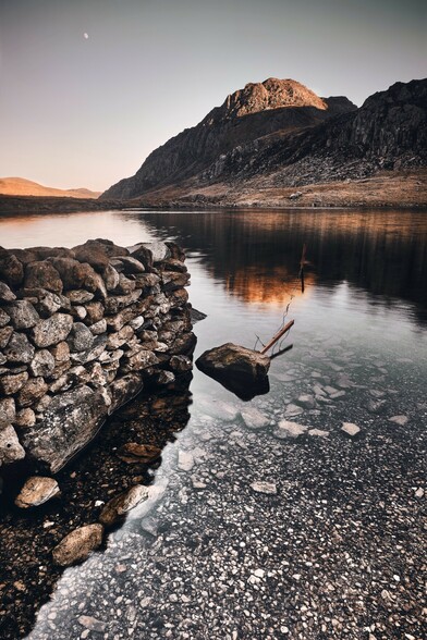 A winter moon rises over Tryfan and Llyn Idwal in Eryri. The light from the setting catches the top of the mountain. The lake is calm. The landscape is silent. 