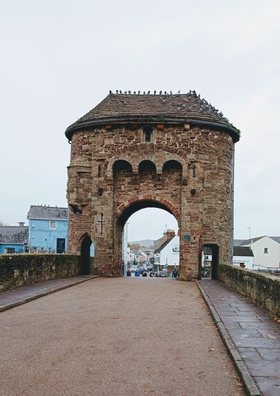 A view through archway of the medieval fortified Monnow bridge. There are a flock of pigeons on the roof of the old jail room above the bridge. Monmouth high street shops can be seen through the archway. 