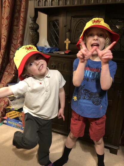 Two young boys in Welsh bucket hats ready for the Wales versus England game. Both are making silly expressions and the older one is holding up his hands in a W for Wales. 