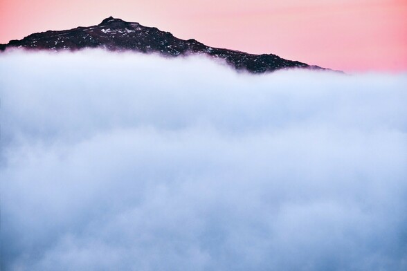 The summit of Yr Wyddfa rises up through a cloud inversion. The peak clear but clouds gather around the lower slopes. 

It is dusk. The sky beyond is pink and purple. It is winter. There is a little snow on the summit. 
