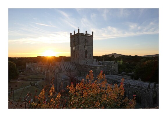The sun setting behind St David's Cathedral in St David's, Pembrokeshire, the smallest city in the UK, with ruins of the Bishop's Palace behind, and sunlit foliage in the foreground