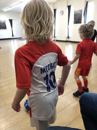 Back of two children. Big brother wears a Croatia kit with Modric on the back. Little brother wears a Wales kit. They are at an indoor football class in a village community centre. 