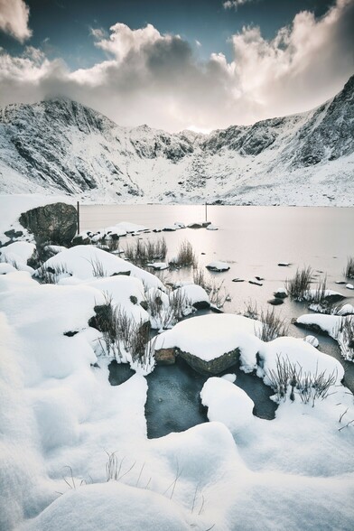 Looking across a frozen Llyn Idwal towards the snow covered mountains beyond. The sky is blue above the clouds. Snow is everywhere. The landscape is white.