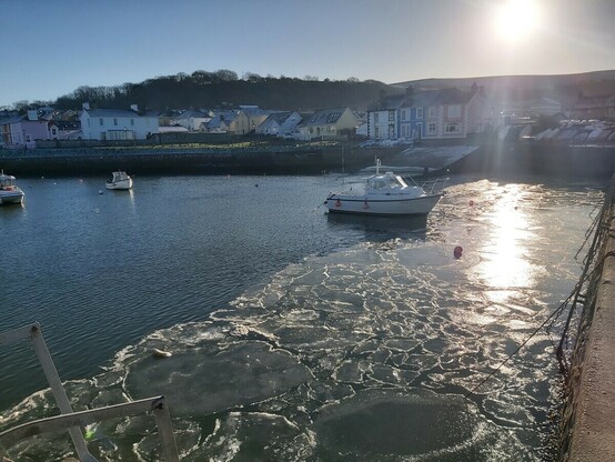 Porthladd a rhew ar wyneb y dŵr. Yr haul yn disgleirio ar yr iâ.
Harbour with ice on the water surface. Sun reflected on ice.