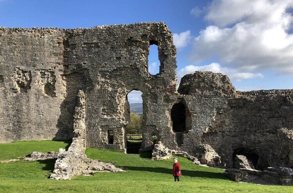 Beautiful, sunny day with fluffy white clouds. A little boy in a red coat stands some distance away. Behind him are the ruins of Denbigh Castle. The grass is bright green. 