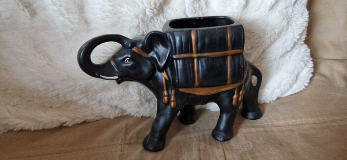 A black elephant ornament/planter carrying a pack on its back. The pack is hollow so could be used for a plant. About 20cm tall. May possibly have originally contained tea. Details are picked out in gold paint.