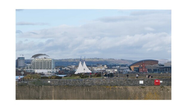 A photo from the Penarth end of Cardiff Bay Barrage, looking across and along the barrage to the Millennium Centre, St David's hotel, Pierhead building and Sendedd at Mermaid Quay, with hills in the far distance. There are quite a lot of people and families walking along the barrage