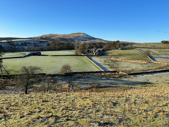 A cold day in the Yorkshire Dales with views across fields with trees and dry stone walls looking to some large hills in the distance with snow and frost on the top