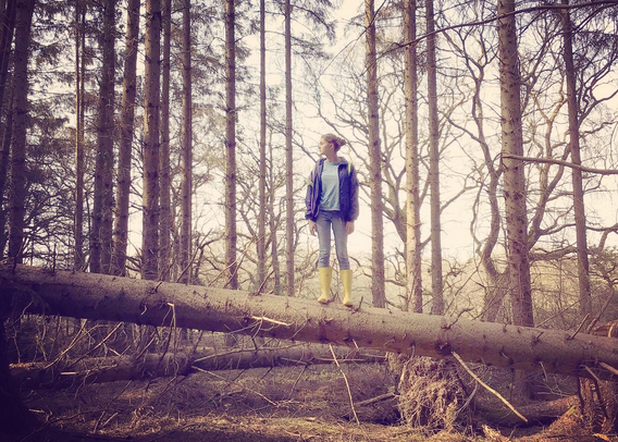 A young adult stands upon a fallen non native tree, within a monoculture commercial forest, they stand square to the viewer, head turned to one side, side profile, looking towards the light.