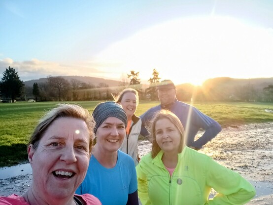 Five friends all in running kit in the centre of the picture. There is a green field behind and hills and trees in the background. The sun is rising over the hill and giving a sun glare effect to the photo. 