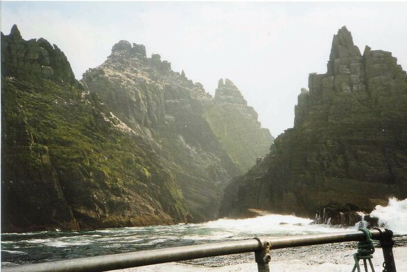 A very rugged island rising out of the Atlantic. The photo is taken from a boat close to the island and a handrail can be seen in the foreground. 4 rocky peaks can be seen and the middle of the island is covered in the small shapes of a multitude of gannets. 