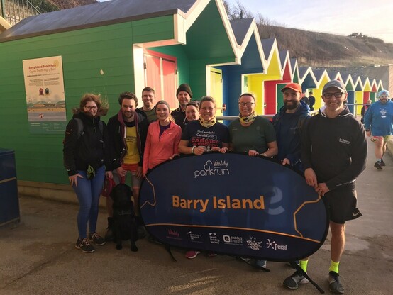 Some San Domenico Club runners around the Barry Island Parkrun sign in front of brightly coloured beach huts on the promenade 