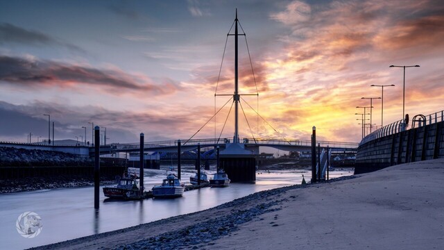 A golden winter sunset. River runningmdiagonally from bottom left to mid right, under a cable stayed foot bridge with golden clouds behind