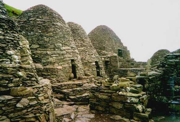 A line of domed monastic cells built from natural stone blocks, similar to that which would be seen in dry stone wall. A single small doorway can be seen at the front of each cell. 