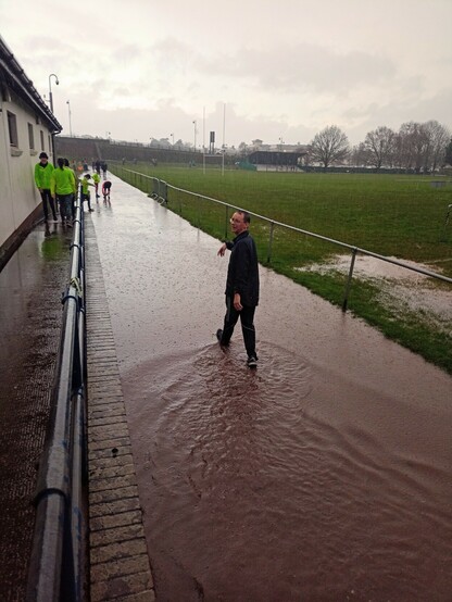A photo of the finish of  a very very wet park run event. There is a man walking through a puddle which is ankle deep turning to face the camera. The puddle covers the path he is walking along and stretches off into the distance. 