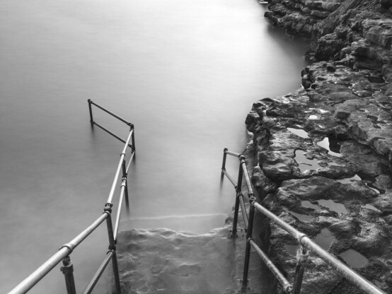 A black and white image of sea steps going into the sea. A long exposure has flattened the water.