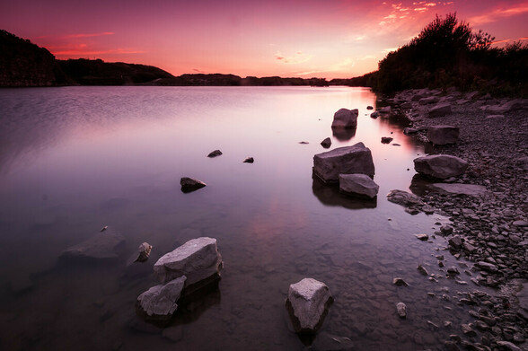 An image of still calm water with foreground rocks. The setting sunset illuminates the scene in a pink hue.
