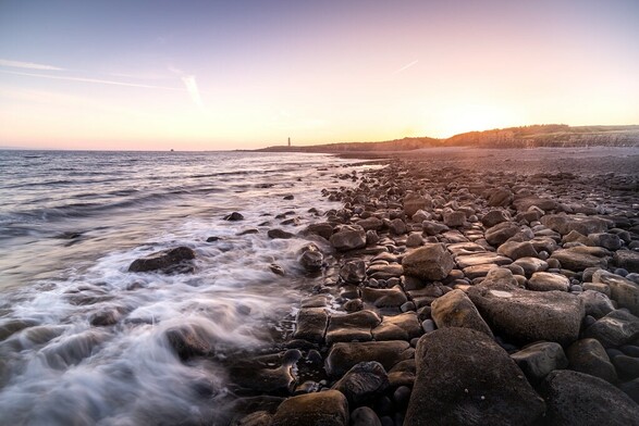 An image of the incoming surf, washing over the foreground of a rocky beach, the sun is just setting.