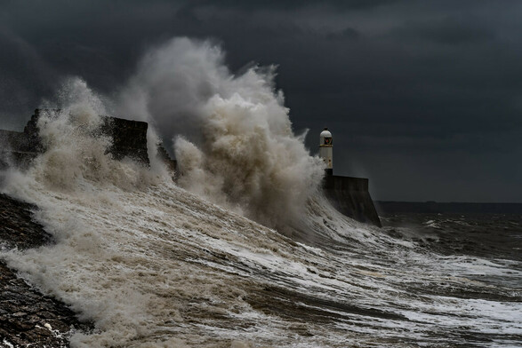 Porthcawl lighthouse being battered by a large wave that's ripping up the breakwater the lighthouse sits on.